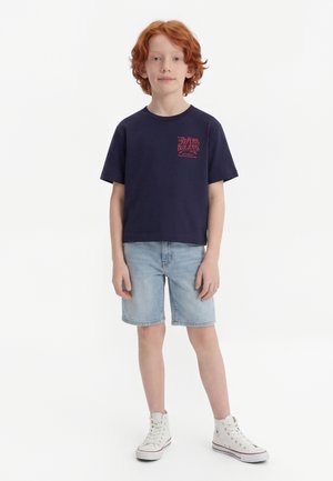 Red-haired boy standing, wearing navy blue T-shirt with red text, light denim shorts, and white high-top sneakers on a white background.