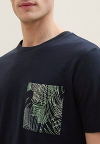 Camiseta de manga corta azul marino con un patrón de hojas de palma en verde y blanco en el bolsillo del pecho. Hecha de material de algodón suave.