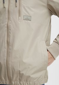 Beige jakke med et glatt, lett stoff, frontglidelås, sidelommer og ribbestrikkede mansjetter. Har et logo-patch på brystet.