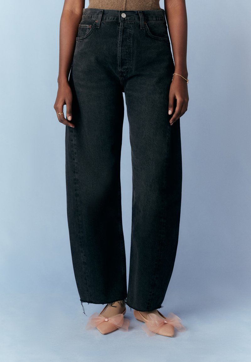 Högmidjade svarta denimbyxor med vida ben och rå nederkant, ihop med ljusrosa lägenheter dekorerade med tyllrosetter och spetsiga tår.