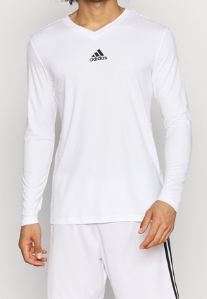 Mężczyzna ubrany w białą koszulkę Adidas z długim rękawem i dekoltem w serek oraz pasujące białe szorty z czarnymi paskami po bokach.