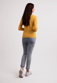 Gelbes langärmeliges Oberteil kombiniert mit hellblauen, leicht verwaschenen Jeans und weißen Sneakers, von hinten betrachtet. Sanfte Stoffstruktur.
