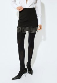 Mini gonna nera con dettagli in frange argentate all'orlo, abbinata a collant opachi neri e scarpe con tacco alto in vernice nera. Camicia bianca semplice con colletto.
