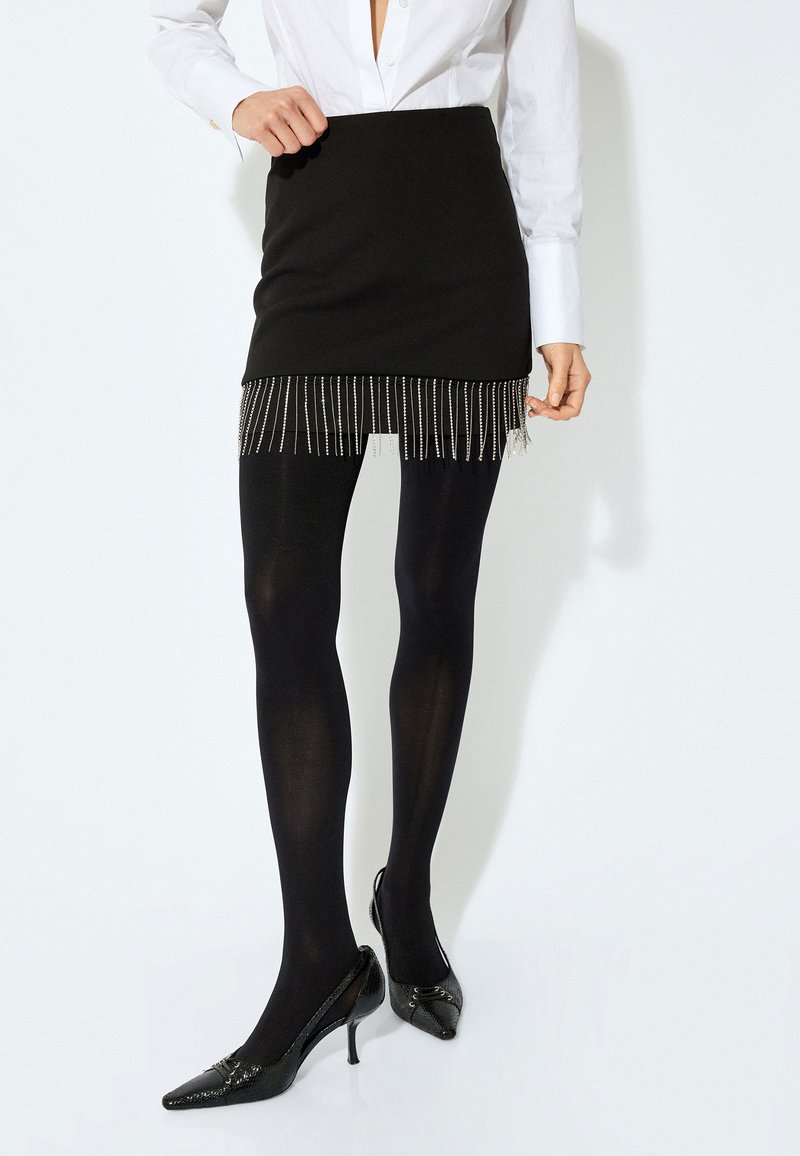Mini gonna nera con dettagli in frange argentate all'orlo, abbinata a collant opachi neri e scarpe con tacco alto in vernice nera. Camicia bianca semplice con colletto.
