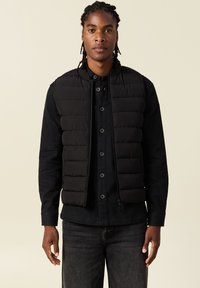 Chaleco acolchado negro con diseño de rombos, cuello alto y cremallera frontal. Lleva puesto sobre una camisa negra de botones, combinada con jeans oscuros.