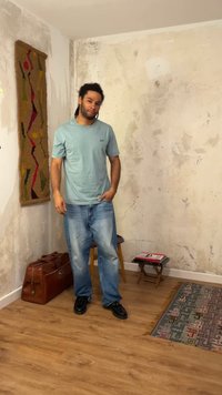 Άνδρας με μπλε μπλουζάκι και τζιν στέκεται σε ένα λιτά διακοσμημένο δωμάτιο με φθαρμένους τοίχους, ξύλινο πάτωμα, ένα χαλί, μια καρέκλα και ένα δερμάτινο τσάντα.