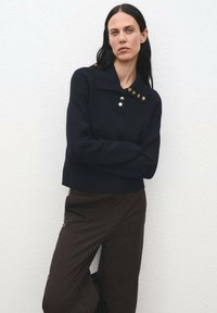 Donna con lunghi capelli scuri che indossa un maglione nero a coste con bottoni dorati e pantaloni marrone scuro, in piedi contro un muro bianco.