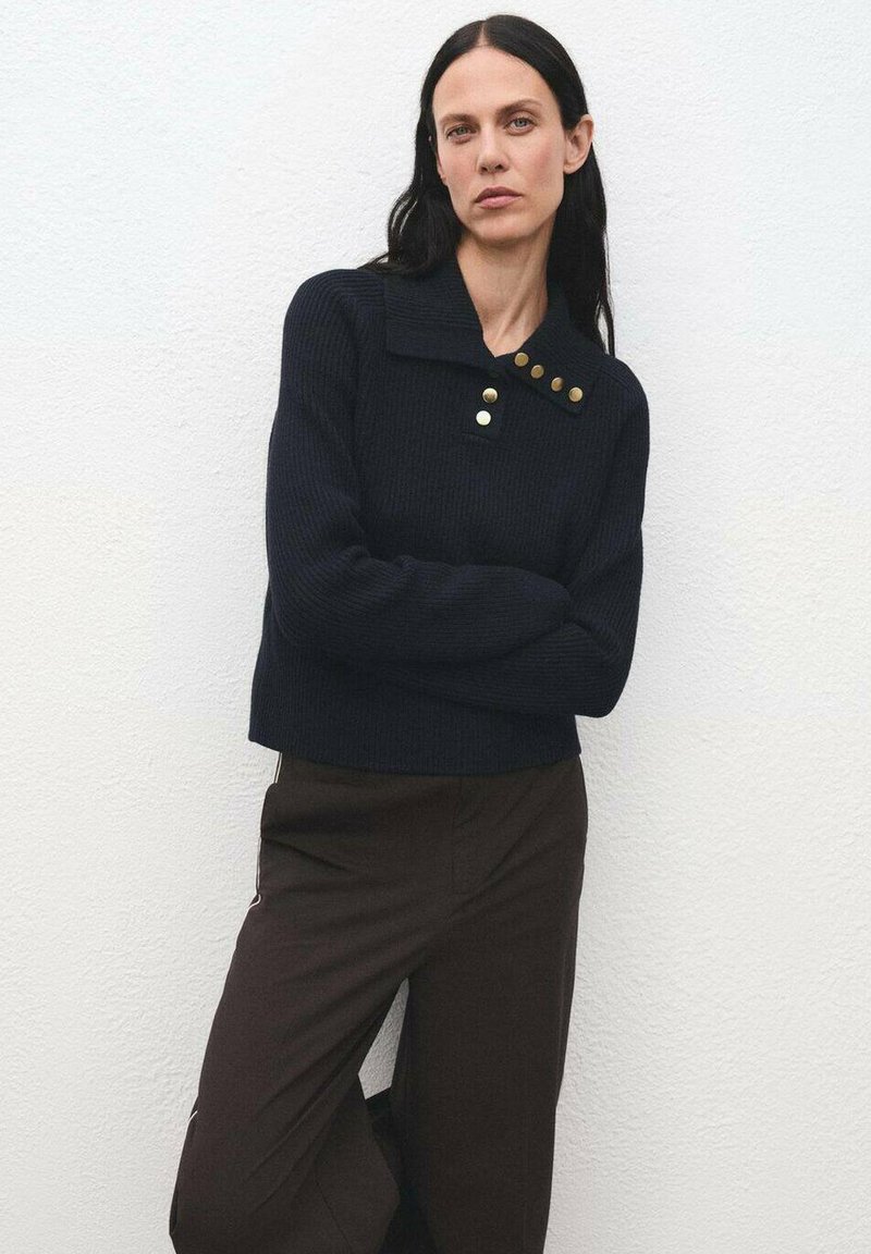 Donna con lunghi capelli scuri che indossa un maglione nero a coste con bottoni dorati e pantaloni marrone scuro, in piedi contro un muro bianco.