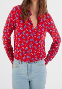 Chemise rouge à boutons avec un motif floral bleu et blanc. Manches longues, col, et tissu léger. Portée avec un jean bleu clair.