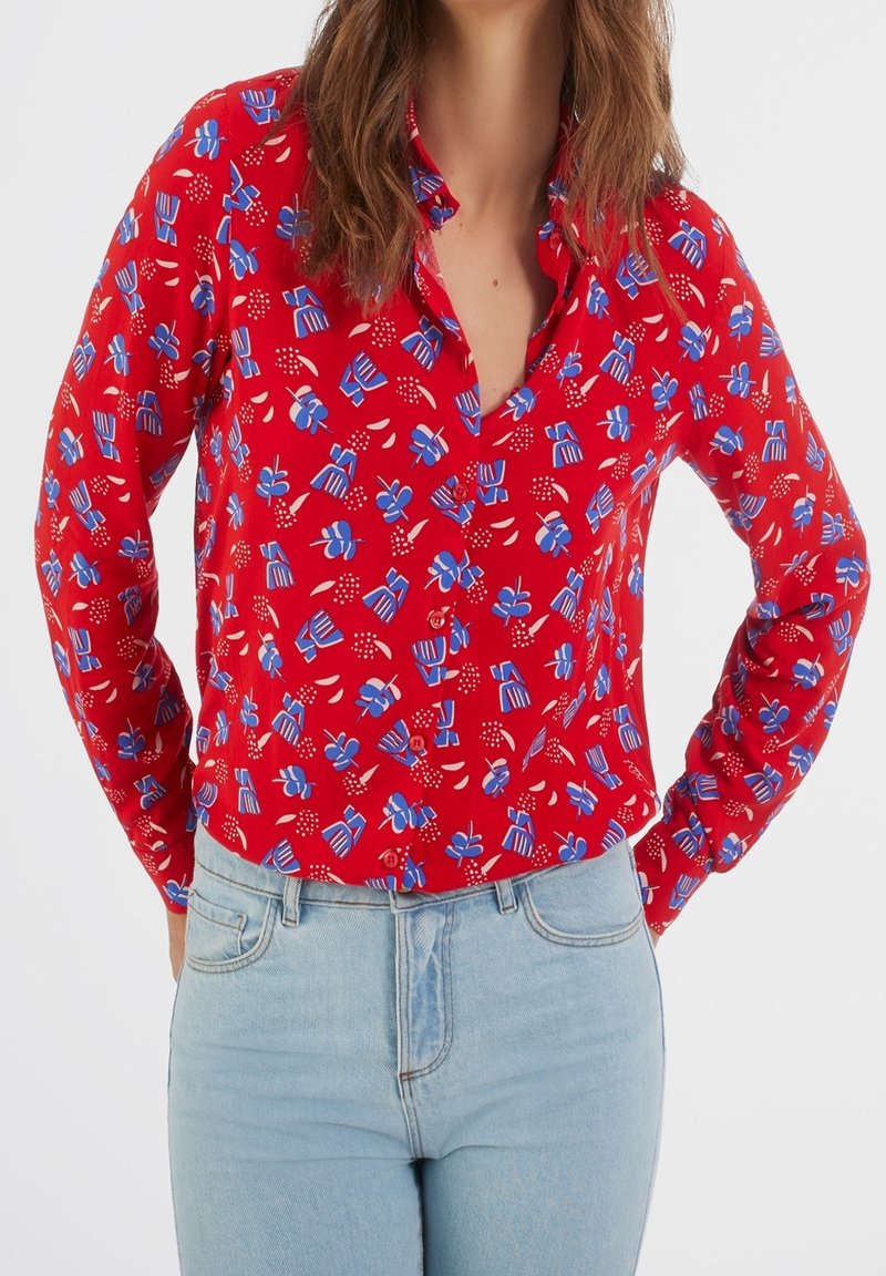 Chemise rouge à boutons avec un motif floral bleu et blanc. Manches longues, col, et tissu léger. Portée avec un jean bleu clair.