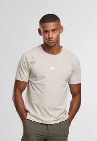 Giovane uomo con capelli corti che indossa una t-shirt attillata beige con un piccolo logo bianco, mani nelle tasche, in piedi contro uno sfondo chiaro e semplice.