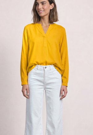 Vrouw draagt een geel hemd met lange mouwen, ingestopt in een witte broek met hoge taille, staand tegen een lichte effen achtergrond.