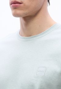 Άνδρας με ανοιχτόχρωμο δέρμα που φοράει ανοιχτογκρι μπλουζάκι με στρογγυλή λαιμόκοψη και ένα μικρό λευκό τετράγωνο λογ ότυπο με την επιγραφή "BOSS" στο στήθος.