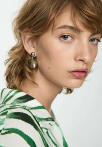 Zilverkleurige oorbellen hebben een bolvormig ontwerp met een gestructureerd oppervlak en een hangend element. Het model draagt een shirt met een groen-witte print.