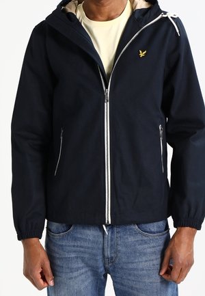 Homme portant une veste à capuche bleu marine avec fermeture éclair devant et poches zippées, associée à un jean bleu clair et un t-shirt beige en dessous.
