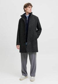 Grå uldblanding frakke med høj krave, frontknapper og to lommer; båret over en navy sweater og en lys blå skjorte, sat sammen med grå bukser.