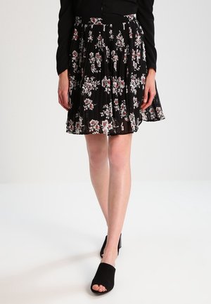 Femme portant une jupe plissée noire avec un motif floral blanc et rouge et des chaussures à talons ouvertes noires sur fond blanc.