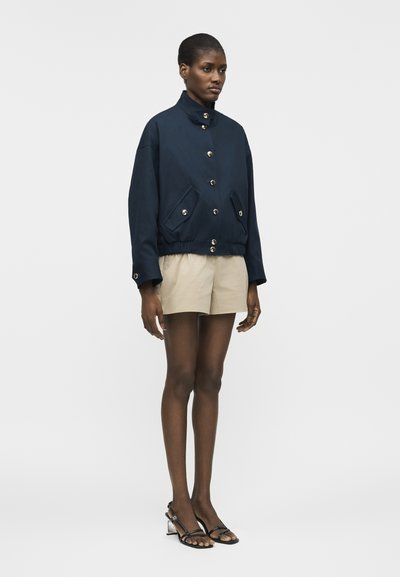 Γυναίκα που φοράει μπλε ναυτικό σακάκι με κουμπιά και τσέπες με καπάκι, μπεζ σορτς και μαύρα σανδάλια με λουράκια και τακούνι, στέκεται μπροστά σε απλό φόντο.