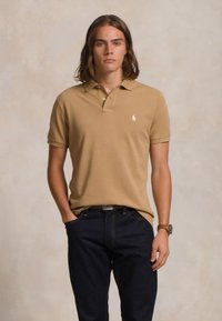 Νεαρός άνδρας με καστανά μαλλιά στο ύψος των ώμων, φορώντας ένα καφετί πόλο μπλουζάκι και σκούρα τζιν, στέκεται με το ένα χέρι στην τσέπη σε ουδέτερο φόντο.