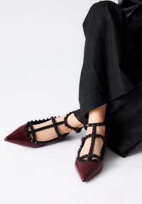 Zapatos de charol rojo con punta afilada y correas negras con tachuelas. El diseño presenta correas entrecruzadas y una silueta elegante.