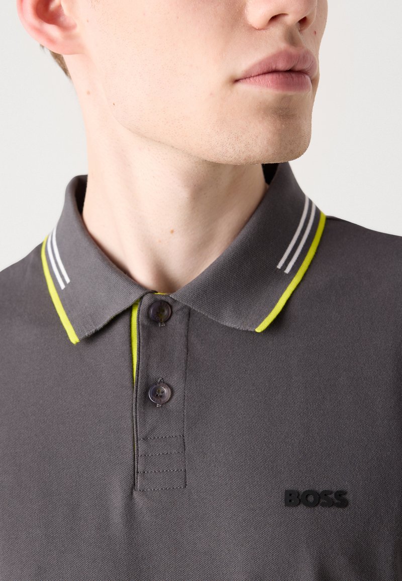 Graues Poloshirt mit einer Knopfleiste mit zwei Knöpfen, ausgestattet mit gelb-weißen gestreiften Kragenakzenten und einem schwarzen "BOSS"-Logo auf der Brust.