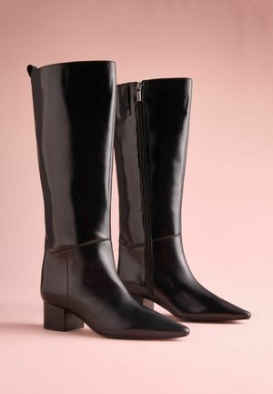 Mustat polvipituiset saappaat, jotka on valmistettu kiiltävästä nahasta. Varustettu teräväkärkisellä muotoilulla, lohkarellasella korolla ja sivuvetoketjulla kiinnitystä varten. Sileä pinta, vähäiset ompeleet.