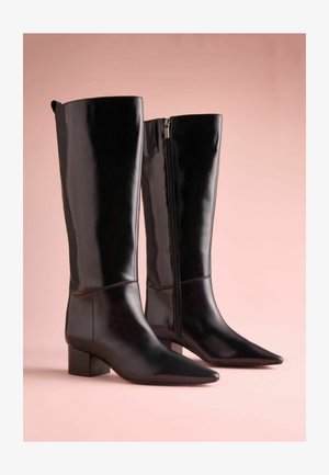 Mustat polvipituiset saappaat, jotka on valmistettu kiiltävästä nahasta. Varustettu teräväkärkisellä muotoilulla, lohkarellasella korolla ja sivuvetoketjulla kiinnitystä varten. Sileä pinta, vähäiset ompeleet.