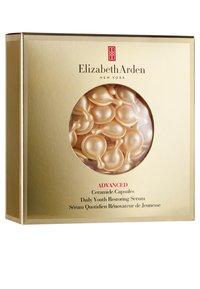 En guldig låda med ett runt fönster som avslöjar mjuka, persikofärgade kapslar. Innehåller text som anger produktnamn och fördelar i rött och svart.