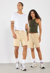 Shorts en coton beige avec taille élastique et cordon de serrage, assortis à un t-shirt blanc à col rond et un haut sans manches vert foncé.