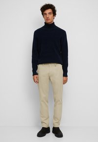 Navy coltrui, beige chino's en donkere instaplaarzen. Model staat tegen een effen achtergrond en toont een casual outfit.