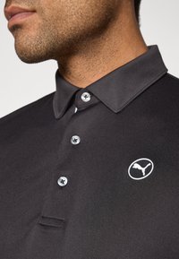 Čierna polokošeľa s golierom, so tromi gombíkmi a logom Puma na ľavej strane. Materiál pôsobí hladko a priedušne.