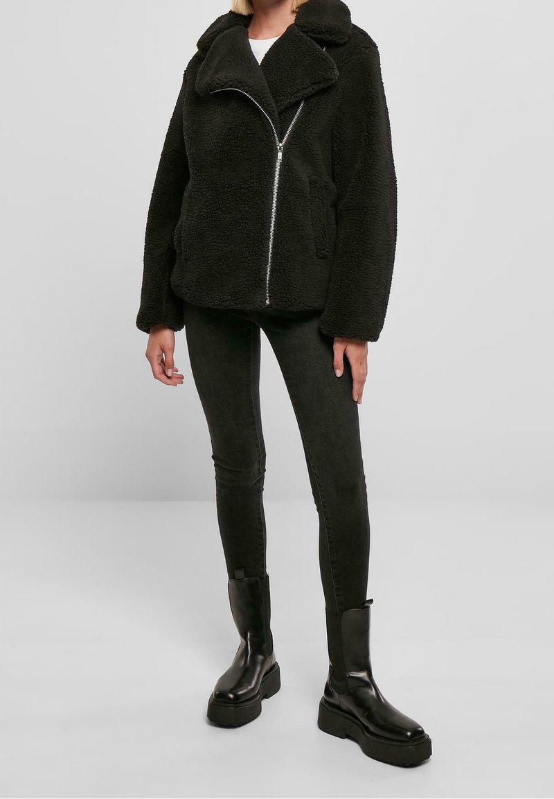 Sort, fluffy sherpa-jakke med bredt revers, frontlynlås og to sidelommer. Kombineret med mørke, stramme jeans og chunky sorte støvler.