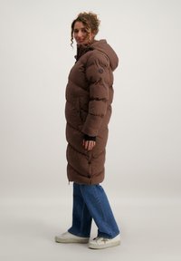 Bruine pufferjas met een capuchon, verticale gestikte ontwerp en drukknoopsluiting. Gemaakt over een zwarte lange mouw top en blauwe wijde jeans.