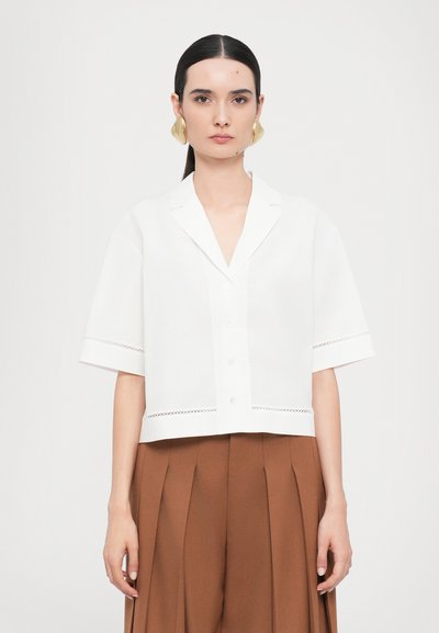 Chemise blanche à manches courtes avec col, fermeture à boutons et dentelle au bas. Associée à un pantalon marron taille haute plissé.