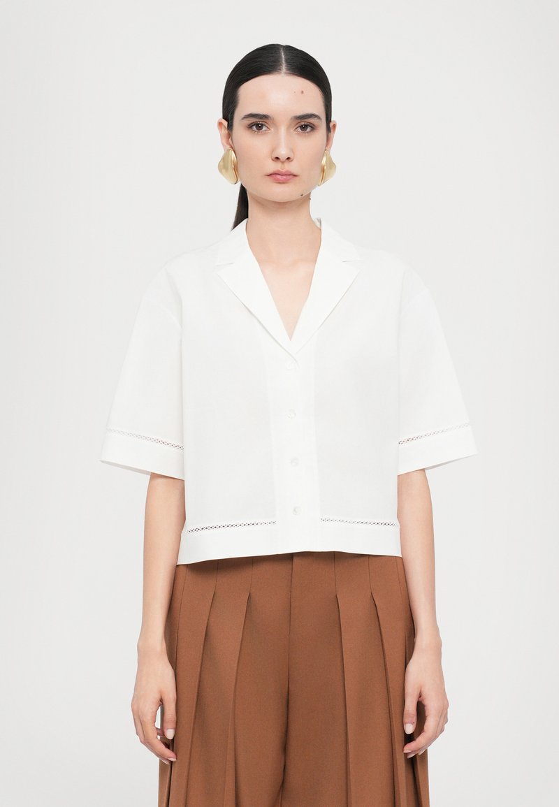 White short-sleeve shirt with a collar, button closure, and lace trim at the hem. Paired with high-waisted, pleated brown trousers.