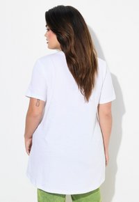 T-shirt en coton blanc à manches courtes et col rond, avec un ourlet arrondi et des fentes sur les côtés, porté sur un pantalon vert en tissu texturé.