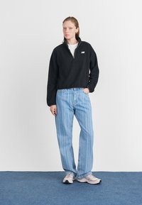 Sudadera de forro polar negra con media cremallera y logo; combinada con jeans de pierna ancha a rayas azules y zapatillas claras. Fondo neutro.