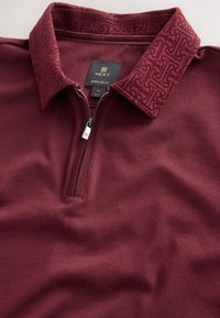 Bordo spalvos polo marškinėliai su užtraukiamu apykakle, turinčiais tekstūruotą bordo raštuotą apykaklę ir lygią medžiagą. Įprasto kirpimo, pažymėta M dydis.