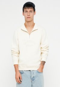 Felpa beige a mezza zip con maniche lunghe, con testo in rilievo "ORIGINALS" sul fronte. Indossata con pantaloni shorts in denim blu chiaro.
