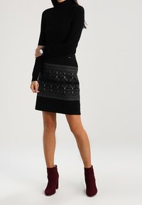 Schwarzes Rollkragenoberteil mit langen Ärmeln, kombiniert mit einem schwarzen Rock mit strukturiertem Muster. Burgunderfarbene Stiefeletten runden das Outfit ab.