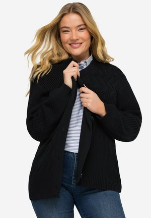 Cárdigan negro de punto con un patrón texturizado, botones frontales y puños acanalados. Combinado con una camisa de rayas con cuello debajo.