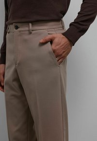 Pantalon beige en tissu doux, avec une poche latérale et une fermeture par bouton. Le design présente une silhouette droite.