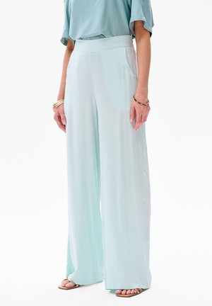 Pantalons larges bleu clair en tissu doux et lisse, avec poches latérales et taille haute, accompagnés de sandales à bout ouvert.