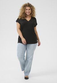 Svart V-ringad t-shirt med korta ärmar, kombinerad med ljusblå jeans och svarta lägenheter. Modellen poserar mot en ljusgrå bakgrund.