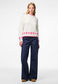 Λευκό πλεκτό πουλόβερ με ροζ σχέδια καρδιάς στο τελείωμα και τις μανσέτες, συνδυασμένο με σκούρες μπλε τζιν παντελόνες φαρδιές και καφέ παπούτσια.