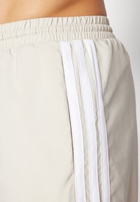 Shorts de sport beiges avec une taille élastique, présentant trois rayures verticales blanches sur le côté et une texture lisse.