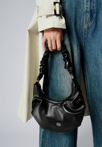 Borsa a mano in pelle nera con manico arricciato, forma curva e dettaglio del logo dorato. Posizionata su uno sfondo chiaro, indossata con denim.