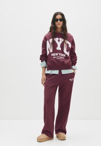 Burgundská mikina s veľkým bielym textom "NYC", doplnená zladenými nohavicami. Prúžkovaná košeľa viditeľná na manžetách. Béžové nazúvacie topánky.