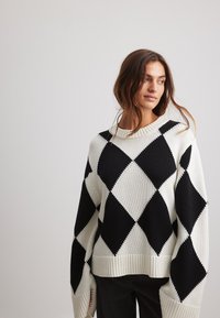 Schwarzes und weißes Argyle-muster Strickpullover aus Strickmaterial. Besitzt einen gerippten Rundhalsausschnitt und weite Ärmel.