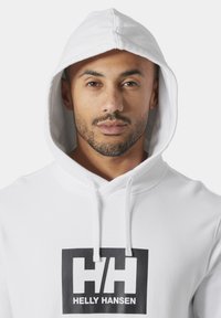 Hombre con barba recortada, vistiendo una sudadera blanca Helly Hansen con capucha puesta, mirando hacia adelante sobre un fondo liso.